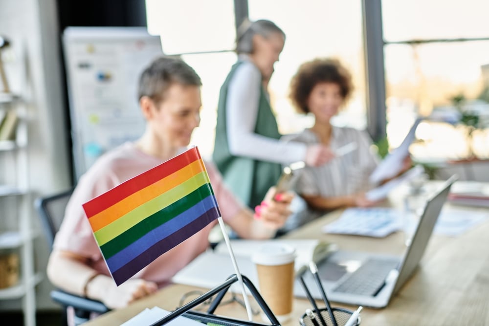 Focus on rainbow flag displayed on office table in front of blurred dverse businesswomen. Foto: Shutterstock