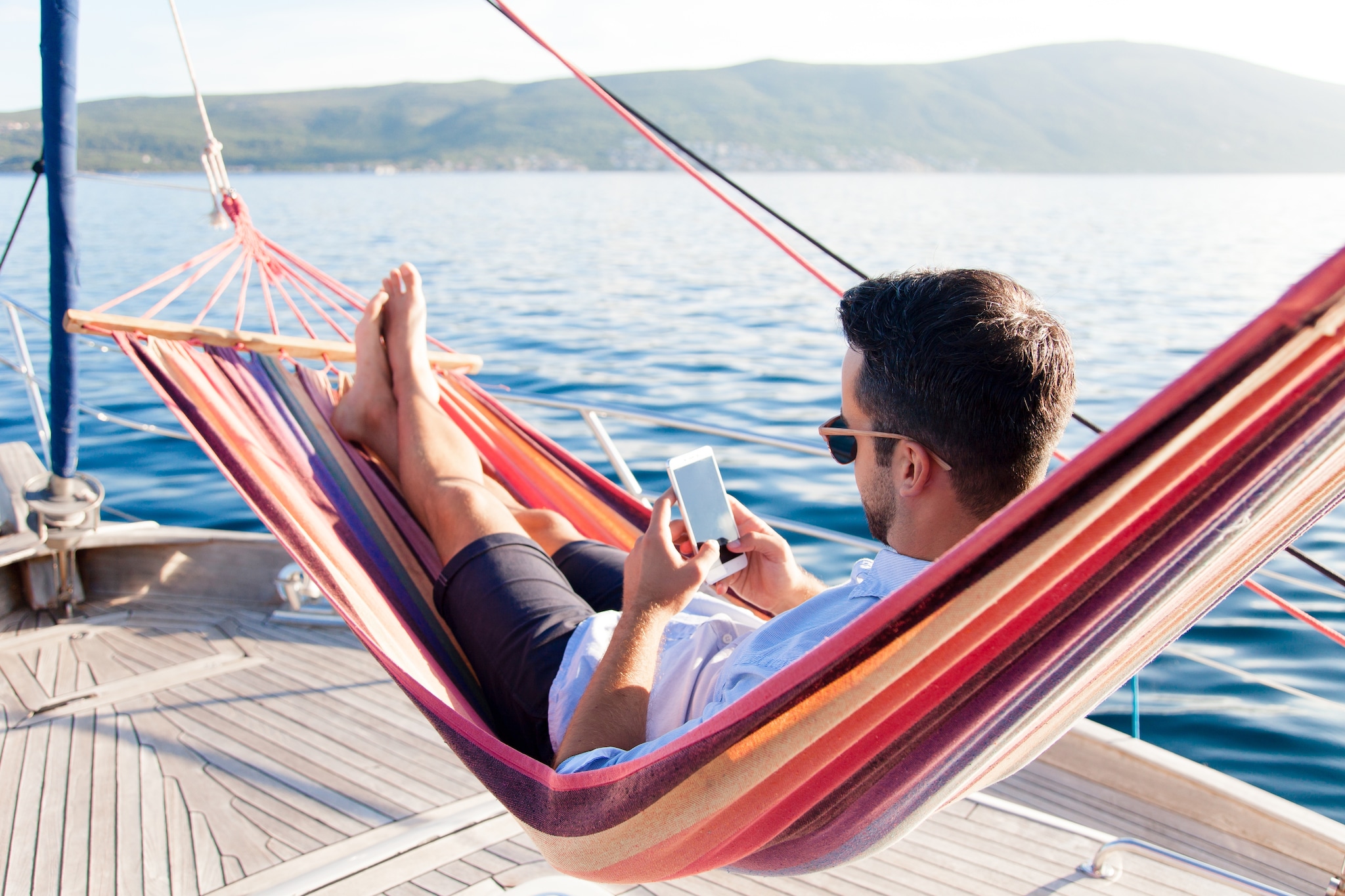 Man with mobile phone relaxing on yacht in hammock. Sea travel on sailboat. Traveler gaming on smartphone in summer vacation. Concept of doomscrolling, digital wellbeing, surfing the Internet, FOMO Shutterstock