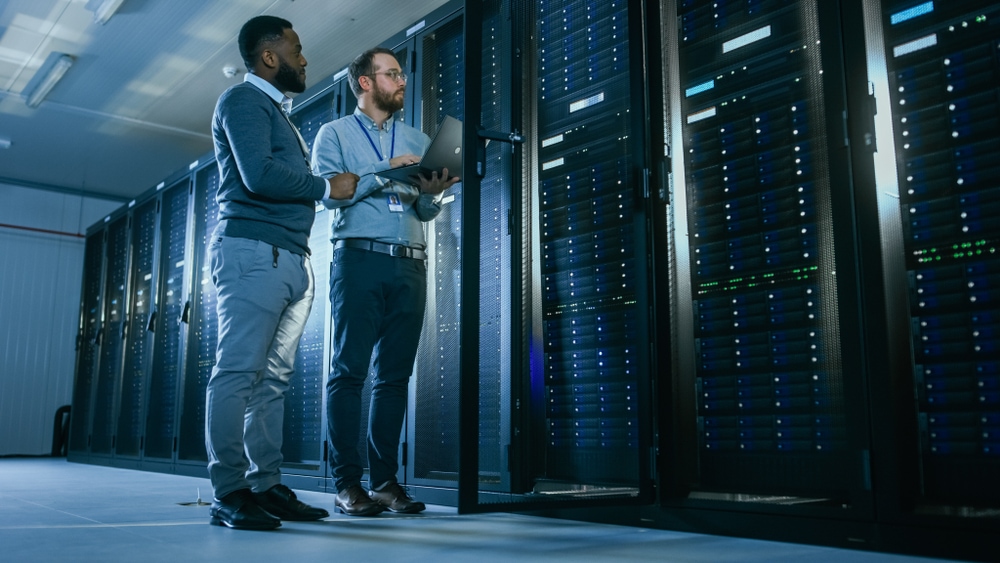 Bearded IT Technician in Glasses with a Laptop Computer and Black Male Engineer Colleague are Talking in Data Center while Working Next to Server Racks. Running Diagnostics or Doing Maintenance Work. De toekomst, ondergronds in Trentino. Foto: Shutterstock.
