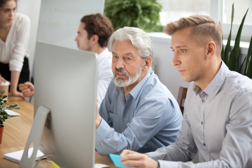 Focused senior and young colleagues discussing online project looking at pc screen, older mentor helps employee explaining computer task, team coworkers talking working together, corporate teamwork Shutterstock