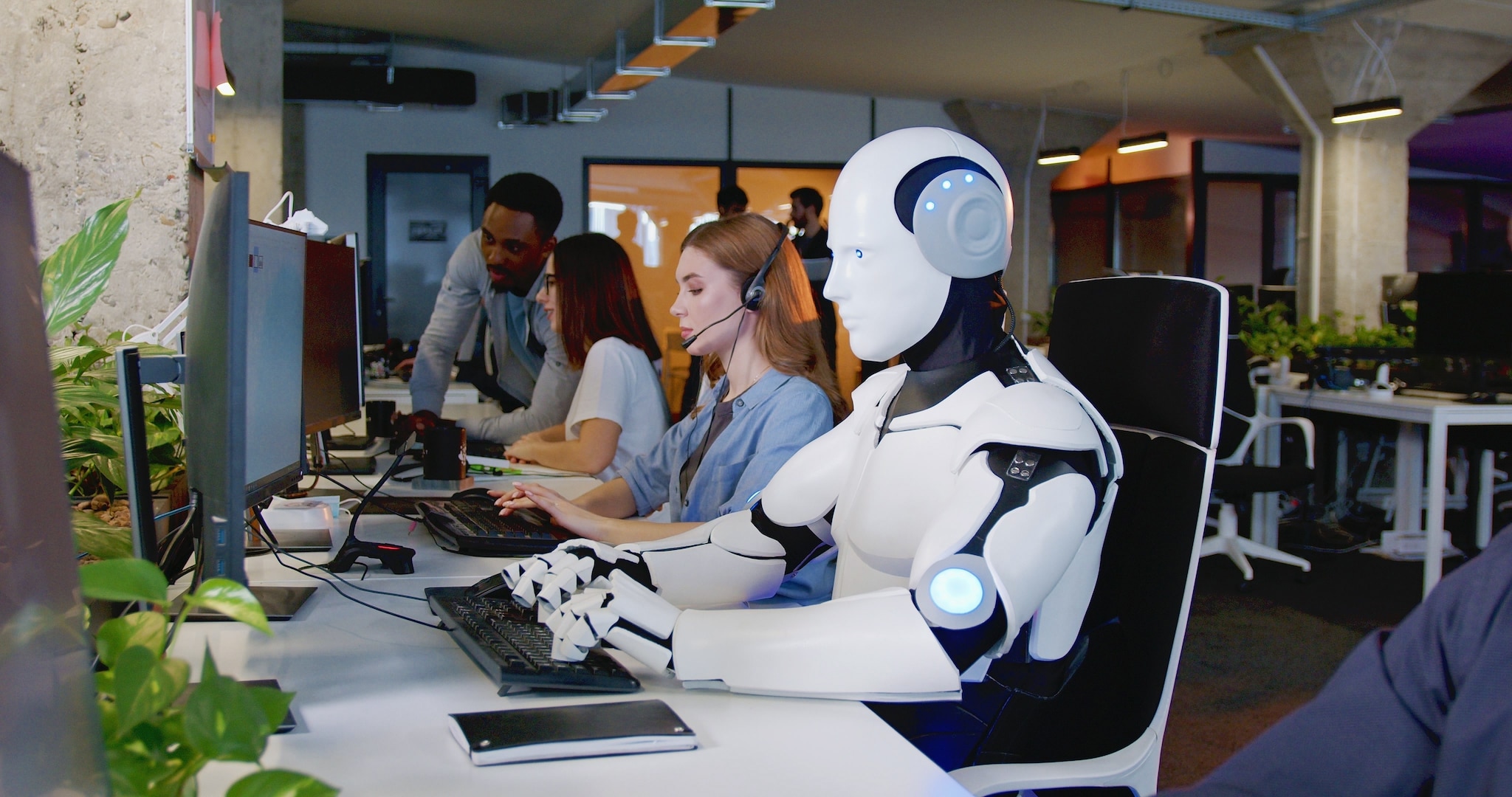 Robot working at computer among people. Maschine typing on keyboard in office. IT team of future. Futuristic worker. Humanoid work at call center. Support job. Selling concept. Technologies. Toekomstmuziek? Ja. Maar wel in de nabije toekomst. Afbeelding: Shutterstock